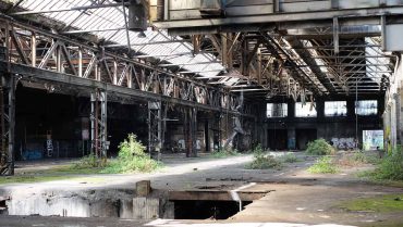 Verlassene Industriehalle mit rostigem Stahlgerüst und zerfallender Struktur, umgeben von Pflanzen, zeigt den Verfall einer alten Fabrik in einer urbanen Umgebung.
