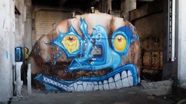 Graffiti-Kunstwerk von Michael Stevens auf einer großen, verrosteten Metallkugel in einer urbanen, verlassenen Industriehalle, mit lebendigen blauen und gelben Farben.