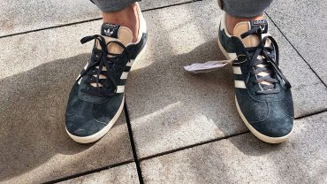 Nahaufnahme von Michael Stevens in schwarzen Adidas Sneakers auf einem Gehweg, Detailaufnahme der Schuhe und des Bodenbelags.