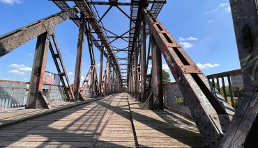 Historische Eisenbahnbrücke aus Stahl mit rustikaler Struktur, Blick entlang der Brücke, Himmel und Wolken im Hintergrund, ideal für Stadt- und Architekturthemen.