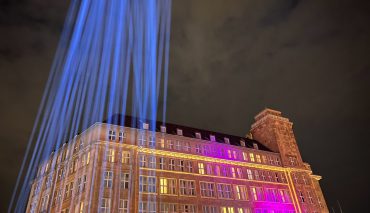 Michael Stevens vor der beeindruckenden Lichtinstallation auf einem historischen Gebäude in Berlin, beleuchtet mit bunten Lichtern und Lichtstrahlen, bei Nacht.