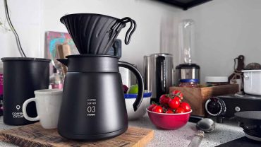 Moderne schwarze Kaffeekanne mit Kaffeefilter auf einer Küchenarbeitsplatte, umgeben von Tassen, einer Schüssel mit Kirschen und Küchenutensilien, stilvolle Küchendekoration.