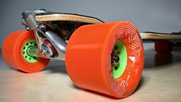 Skateboard mit orange Rollen und grünem Achsenschutz, ideal für Freizeit und Tricks, auf glattem Boden liegend.