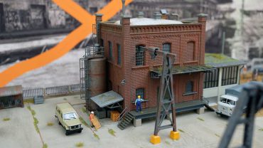Modell eines Industriegebäudes mit Figuren, Fahrzeugen und elektrischen Anlagen, detailreiche Miniaturdarstellung eines industriellen Szenarios.